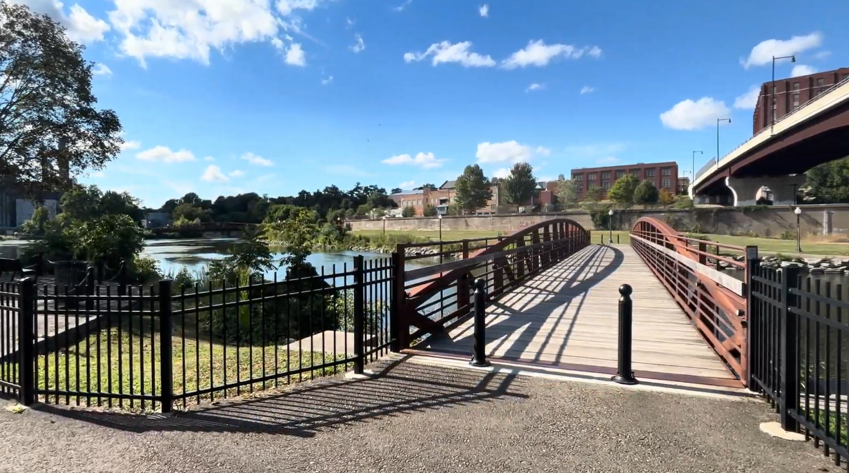 Curved pedestrian bridge with red metal railings crossing the Chadakoin River on a sunny day, with trees, waterfront buildings, and a highway bridge in the background.