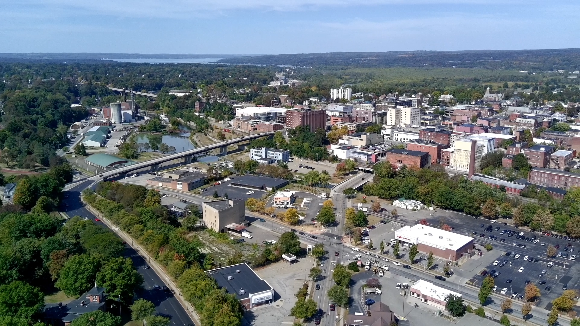 Aerial view of downtown Jamestown with mixed-use buildings, roads, parking lots, and surrounding tree-covered areas extending toward a Chautauqua Lake in the distance.